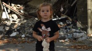 Pigen Lilje står med en bamse i hånden foran murbrokker i Ukraine.